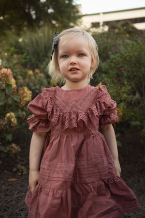 A toddler wearing a dusty rose ruffled dress stands in a garden at sunset.