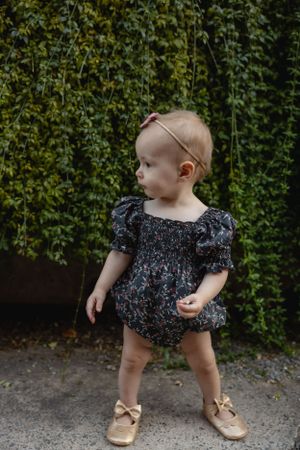 A small child in a floral romper and gold shoes stands against a vine-covered wall.
