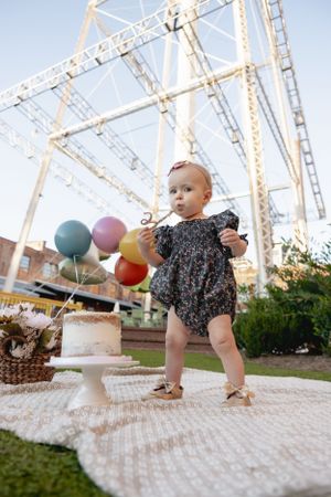 A child in a floral romper stands near colorful balloons and a white cake outdoors.