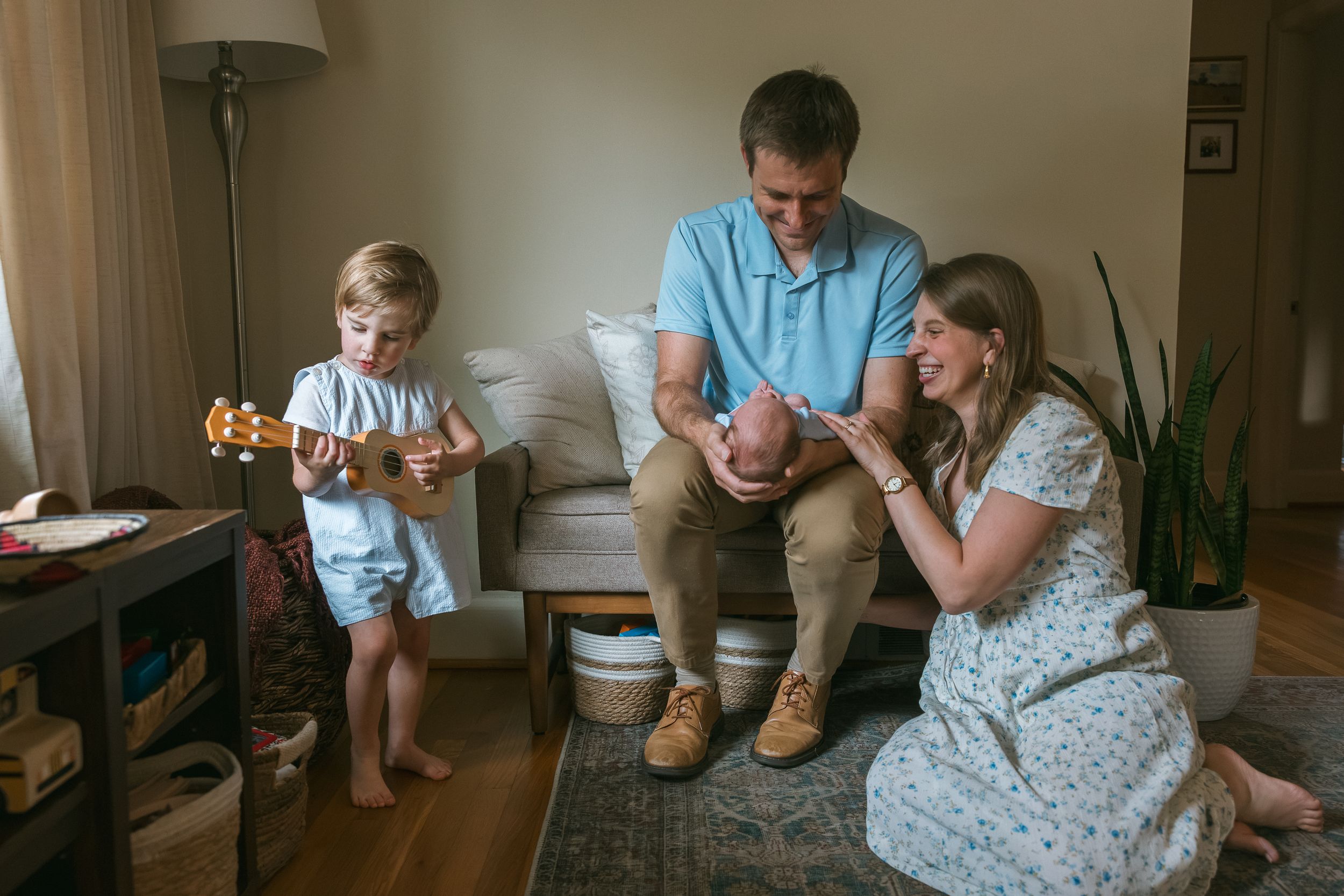 Family at home with toddler playing ukulele while parents admire newborn baby in cozy living room.