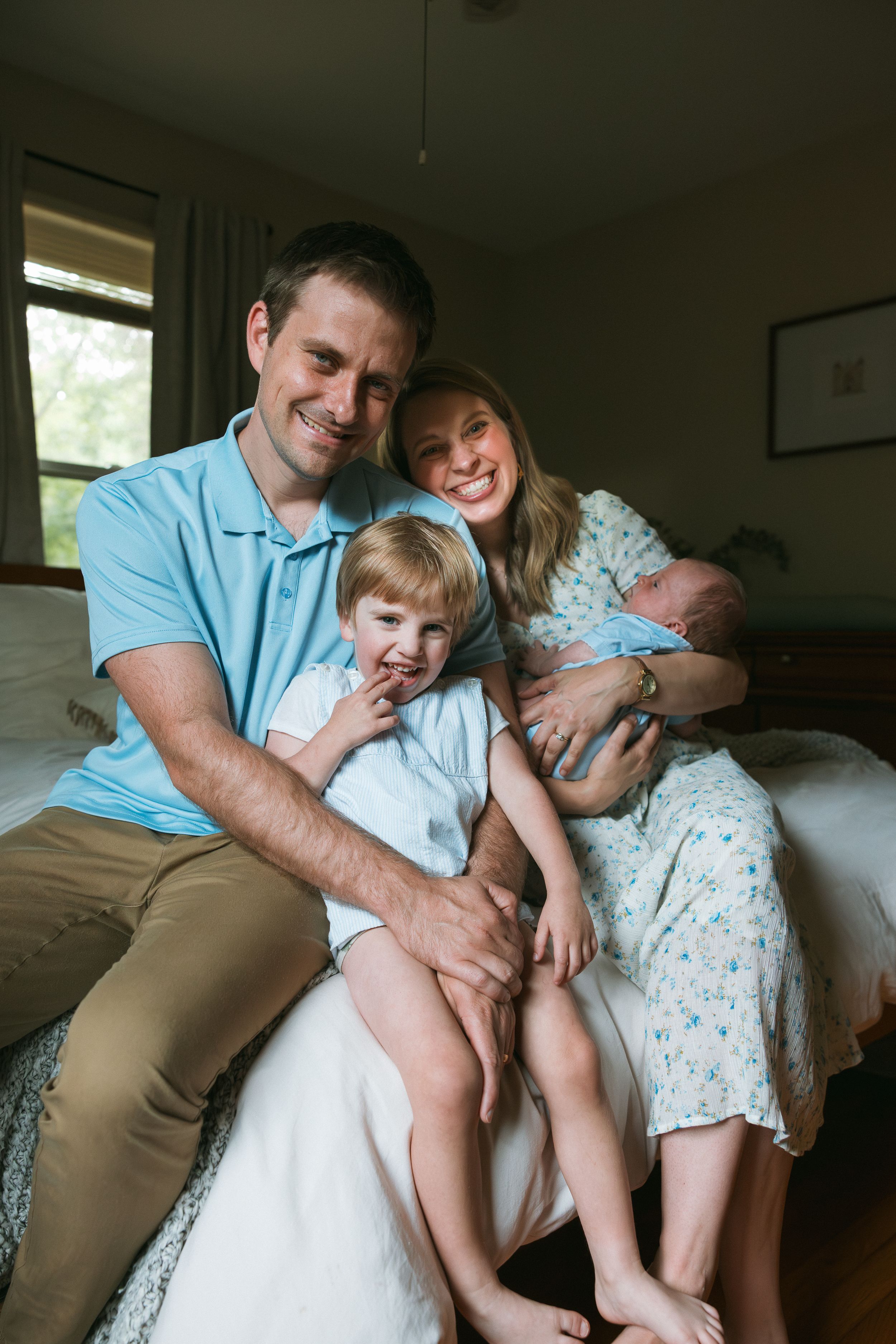 Happy family of four with parents smiling, toddler, and newborn baby sitting together on a bed in a cozy bedroom.