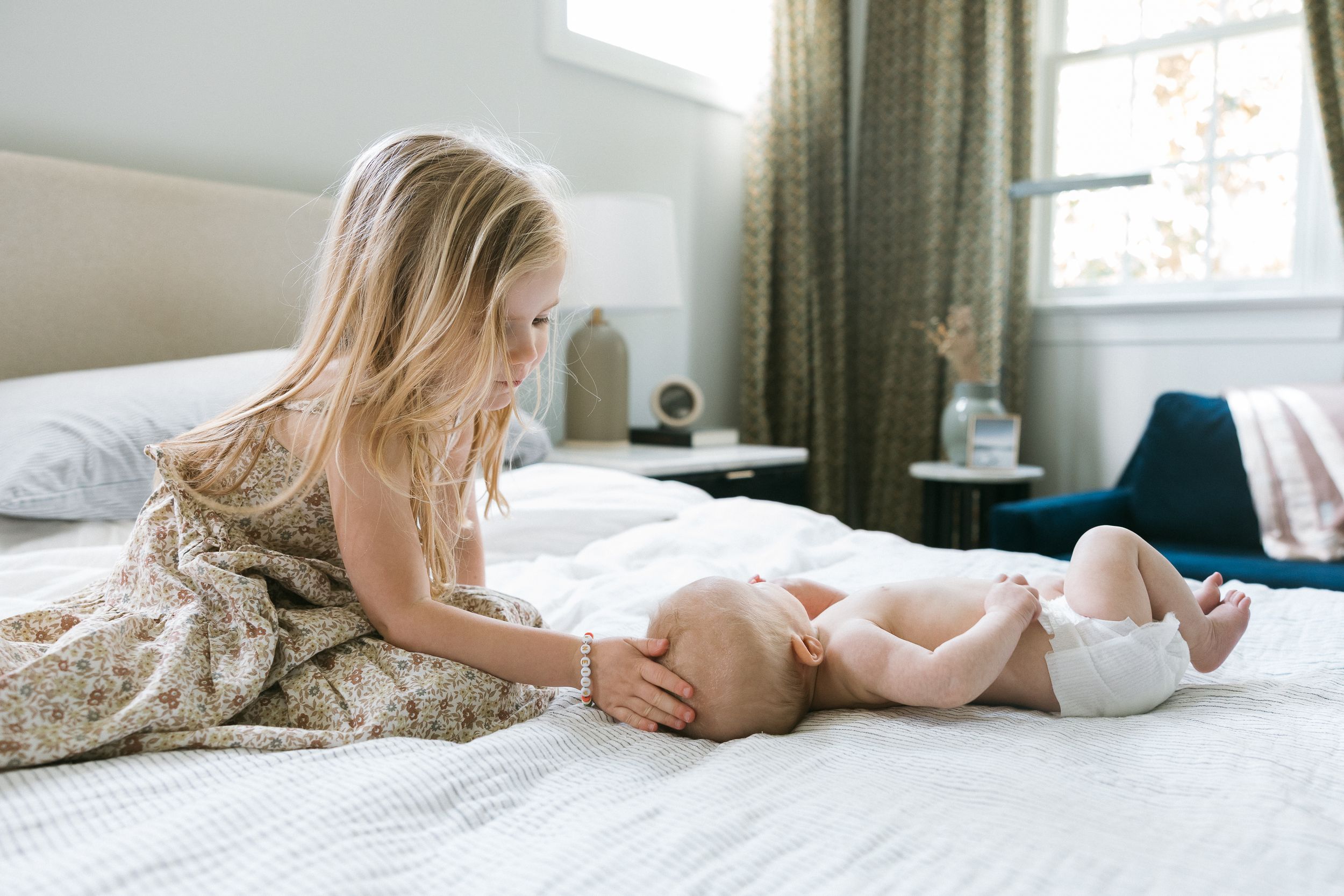 Young girl in floral dress sitting on white bed interacting with a newborn baby in a diaper.
