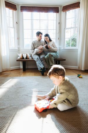 A young family relaxes at home, parents sitting on a bench with a newborn while a toddler plays with toys on a sunlit rug.