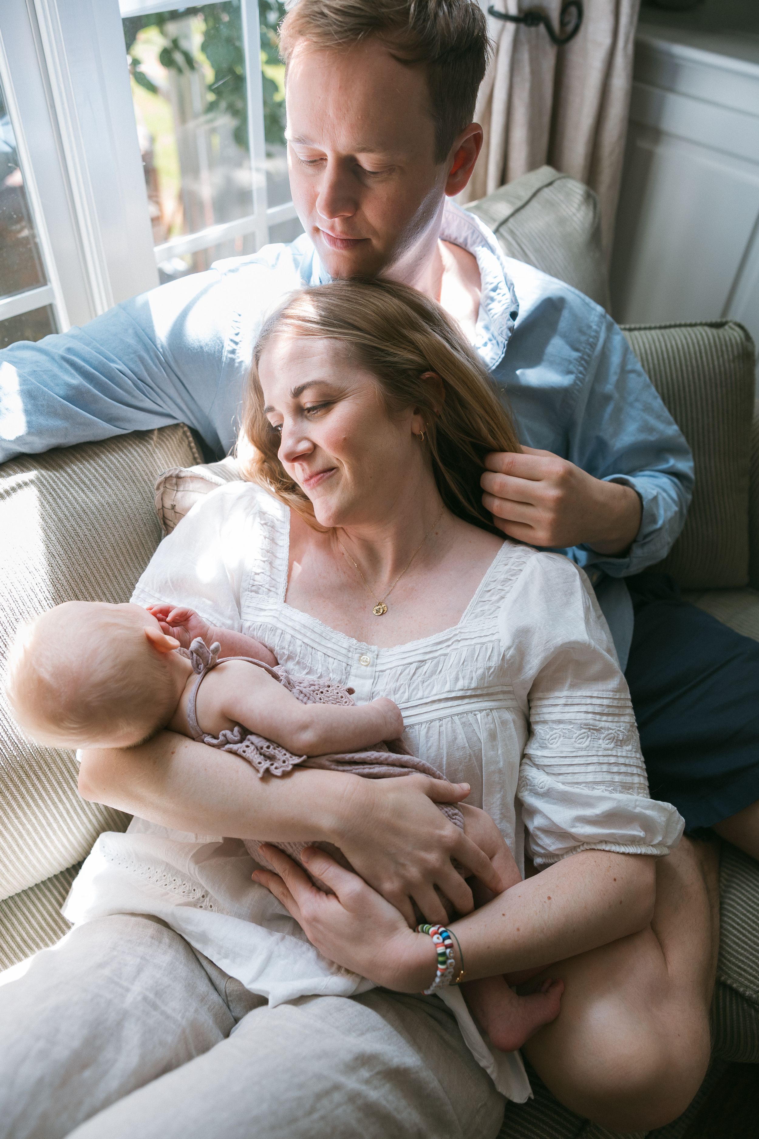 Loving couple sitting on a couch, mother in white dress cradling newborn baby while father gently embraces her.