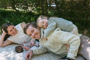 Happy family lying on a blanket outdoors, father smiling with two daughters and a newborn baby in a lush green park.