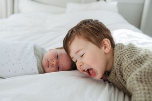 Adorable toddler yawning next to a sleeping newborn baby wrapped in white swaddle on a white bed.