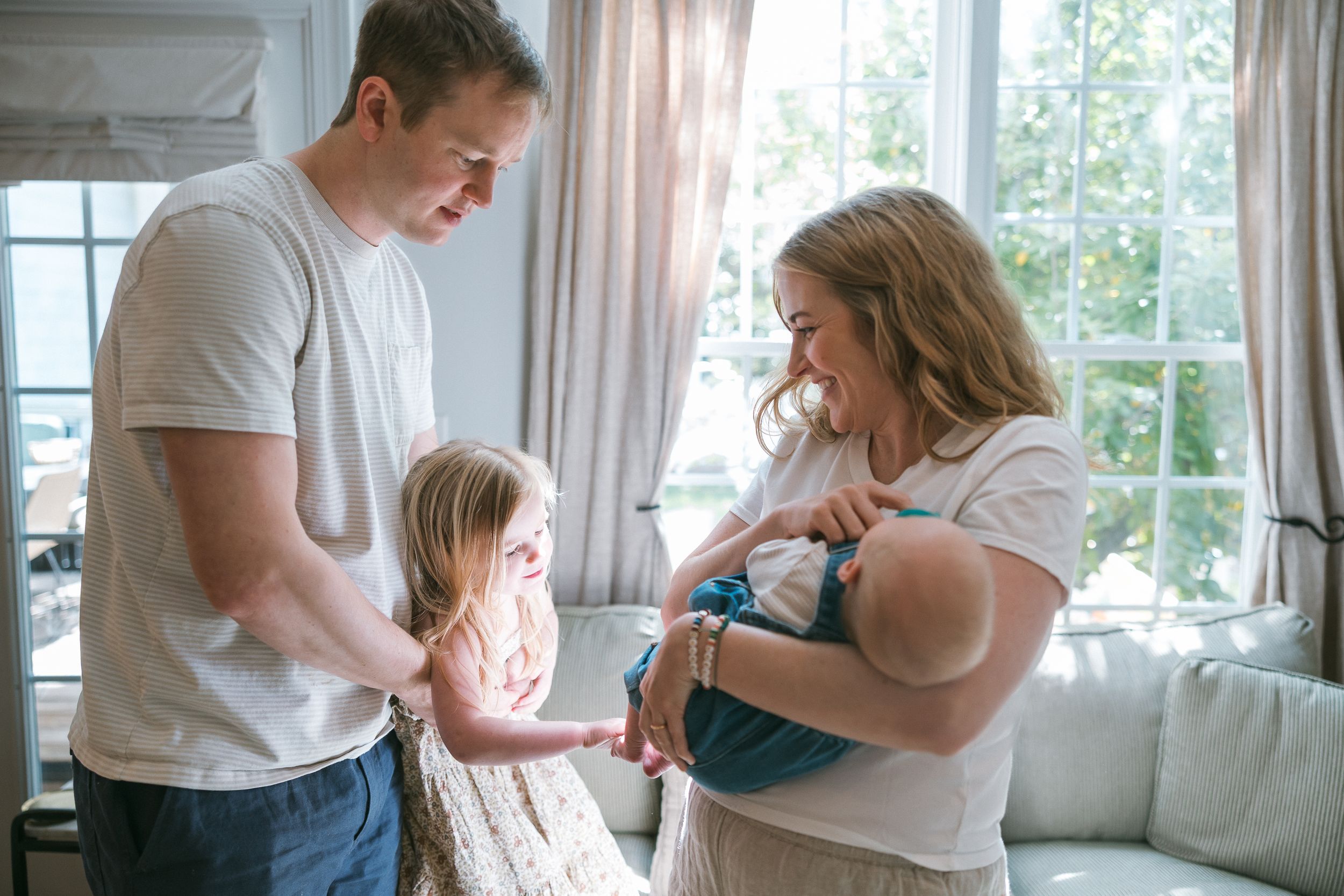 A young family with a toddler girl gathers in a bright living room as the mother holds a newborn baby.