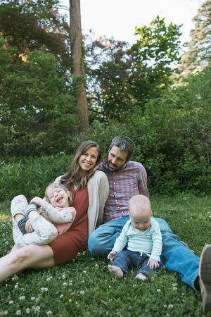 A happy family of four sits on green grass outdoors, with two parents smiling and two young children laughing in a park.
