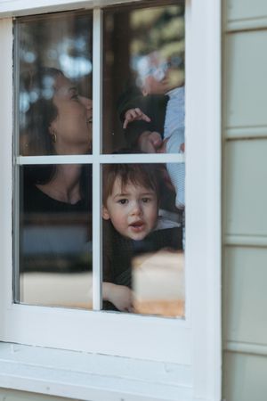A toddler and mother looking through a white-framed window from inside their home.