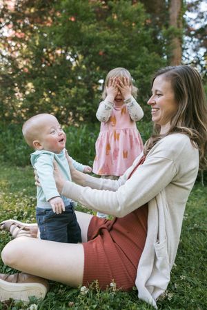A smiling mother sits on grass playing with her baby and toddler daughter in a lush green park.