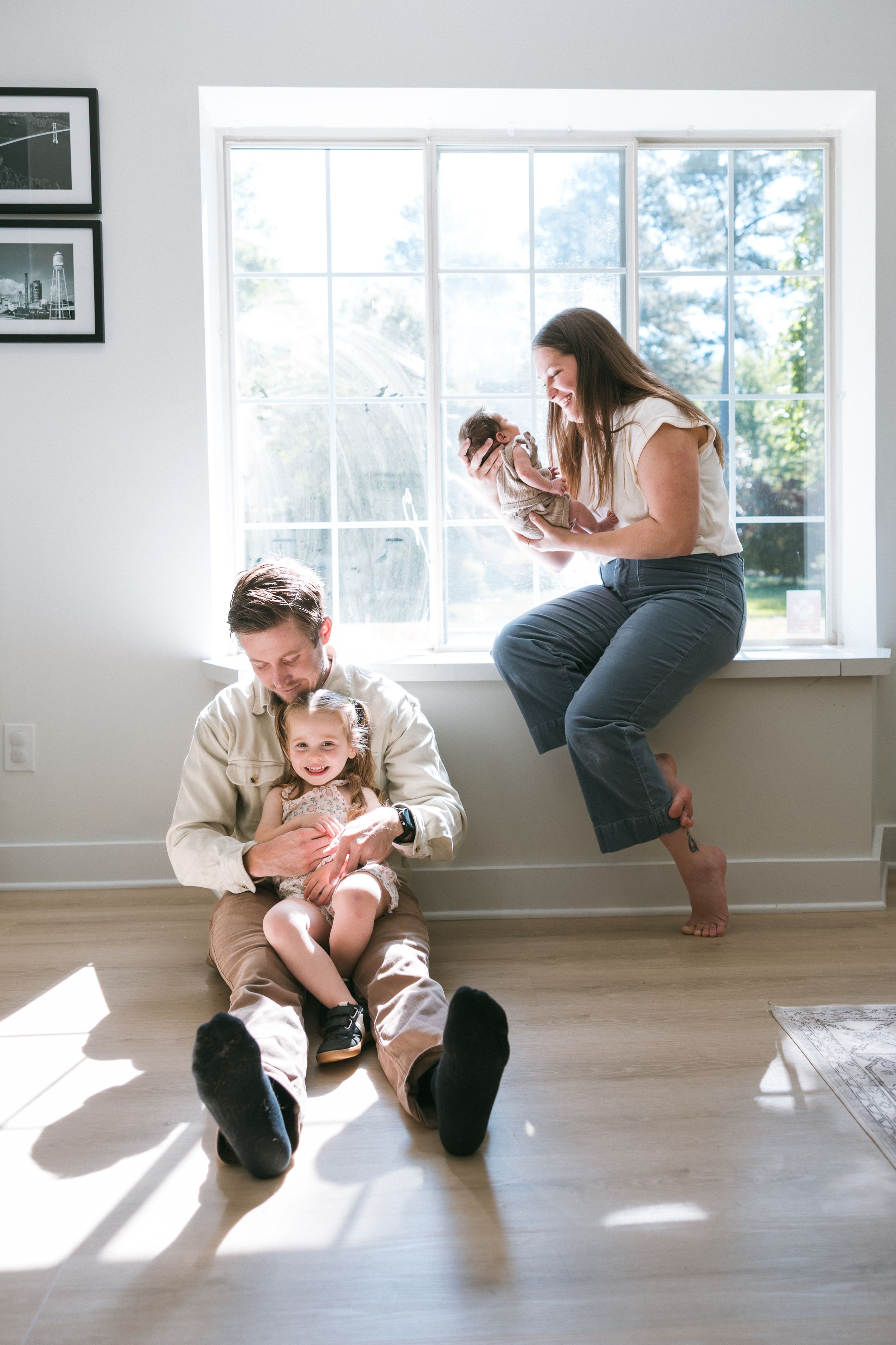 Family of four at home, dad sitting on floor with toddler, mom perched on windowsill holding newborn baby.