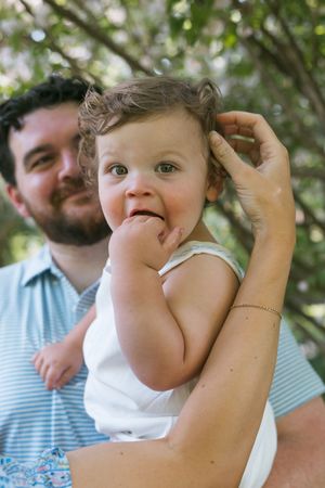 A man smiling while holding a cute baby with curly hair outdoors near green trees.