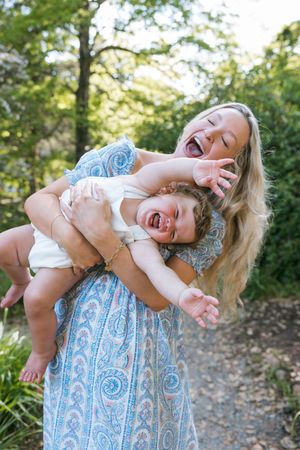 A joyful mother in a blue dress playfully holds her laughing toddler outdoors on a garden path.