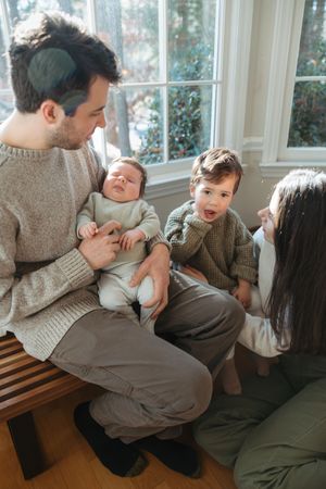 A family of four sits by a window, father holding a newborn while mother interacts with their excited toddler.