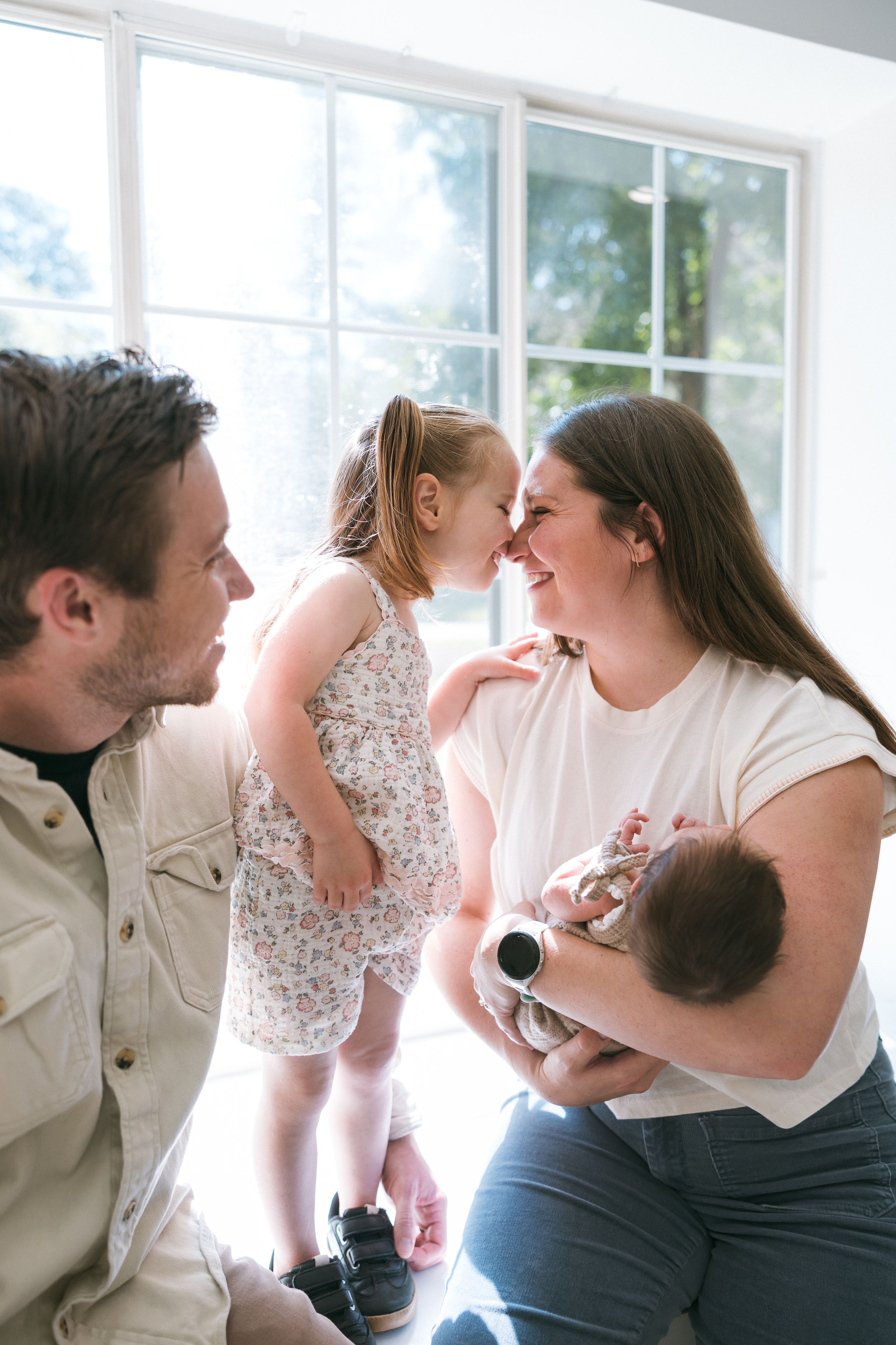 A young family with a toddler girl and newborn baby sharing a tender moment together indoors near a bright window.