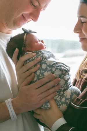 New parents lovingly gaze at their swaddled newborn baby girl wearing a floral wrap and bow headband.