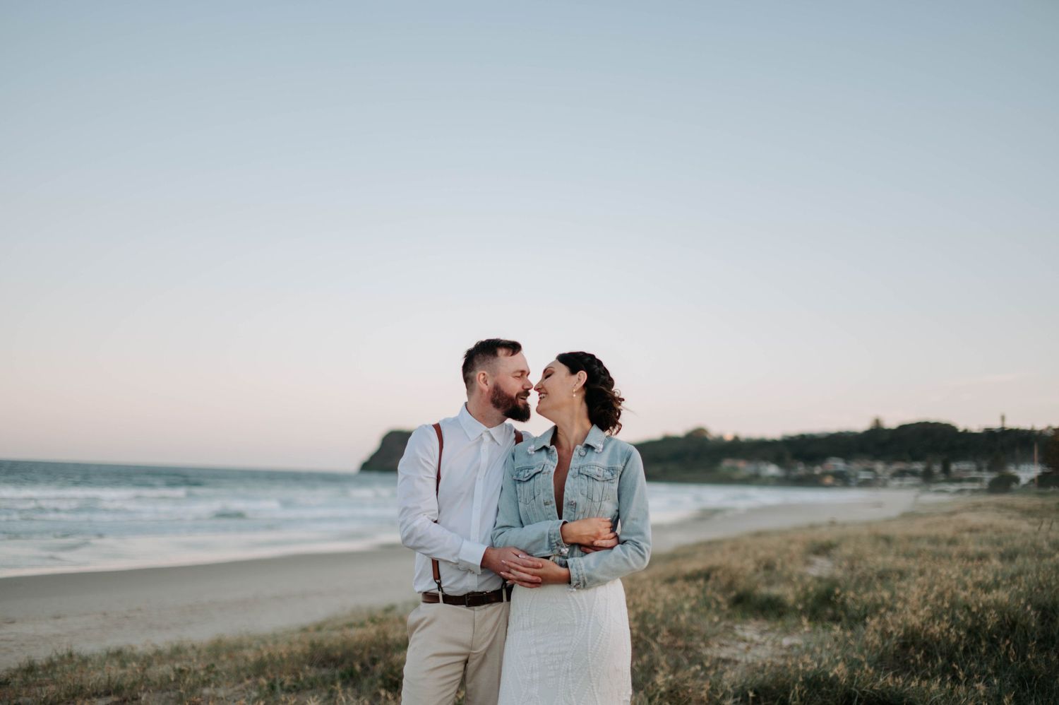 Romantic coastal wedding portrait of couple embracing at sunset on beach.