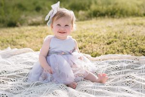 Baby wearing light purple dress and bow sits smiling on white blanket in sunny outdoor setting.