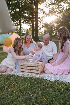 Family having outdoor picnic celebration with balloons and wooden crate on white lace blanket at sunset in park.