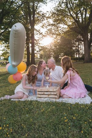 A group sits on a picnic blanket in a sunlit park with colorful balloons and a wooden crate during golden hour.