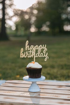 A chocolate cupcake with glittery gold 'happy birthday' topper sits on a light blue cake stand outdoors.