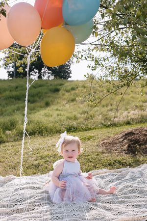 A small child in a tulle dress sits on a lace blanket outdoors with colorful balloons floating above.