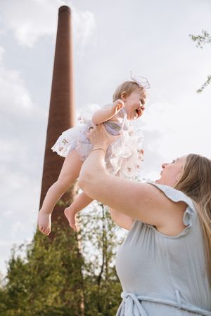 A joyful moment captured against an industrial brick chimney as a person in gray lifts a laughing baby in a white dress.
