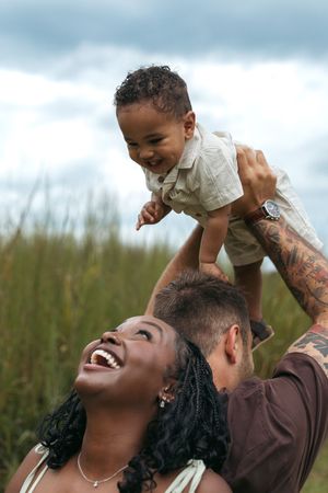 A joyful interracial family plays outdoors, father lifting smiling toddler overhead while mother laughs in a grassy field.