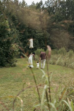 Family of three walking in lush green meadow, parents holding toddler's hands with number 1 balloon on first birthday.