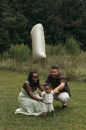 Interracial family celebrating baby's first birthday with a number one balloon on green grass in a park.