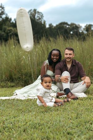 Interracial family sitting on grass in a park, smiling with their baby and a white balloon floating nearby.