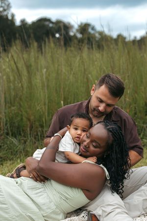 Interracial family sitting in a grassy field, parents lovingly embracing their baby outdoors.