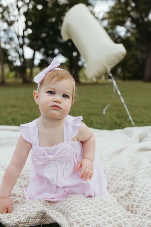 Small baby wearing pink dress and bow sits on blanket outdoors with white balloon in background.