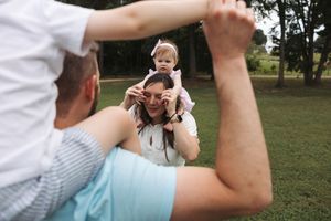 A family plays together outdoors in a park, with a small child being lifted up playfully on someone's shoulders.