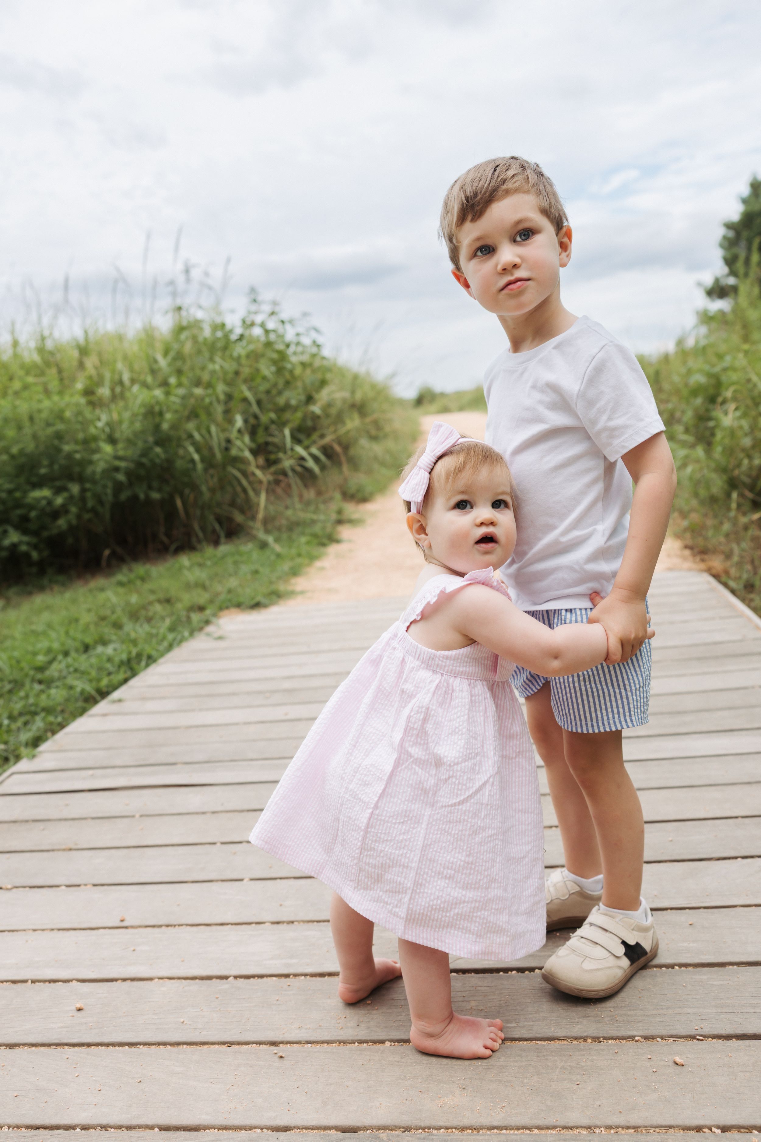 Two young children interact while walking on a wooden boardwalk path through natural scenery on a bright summer day.