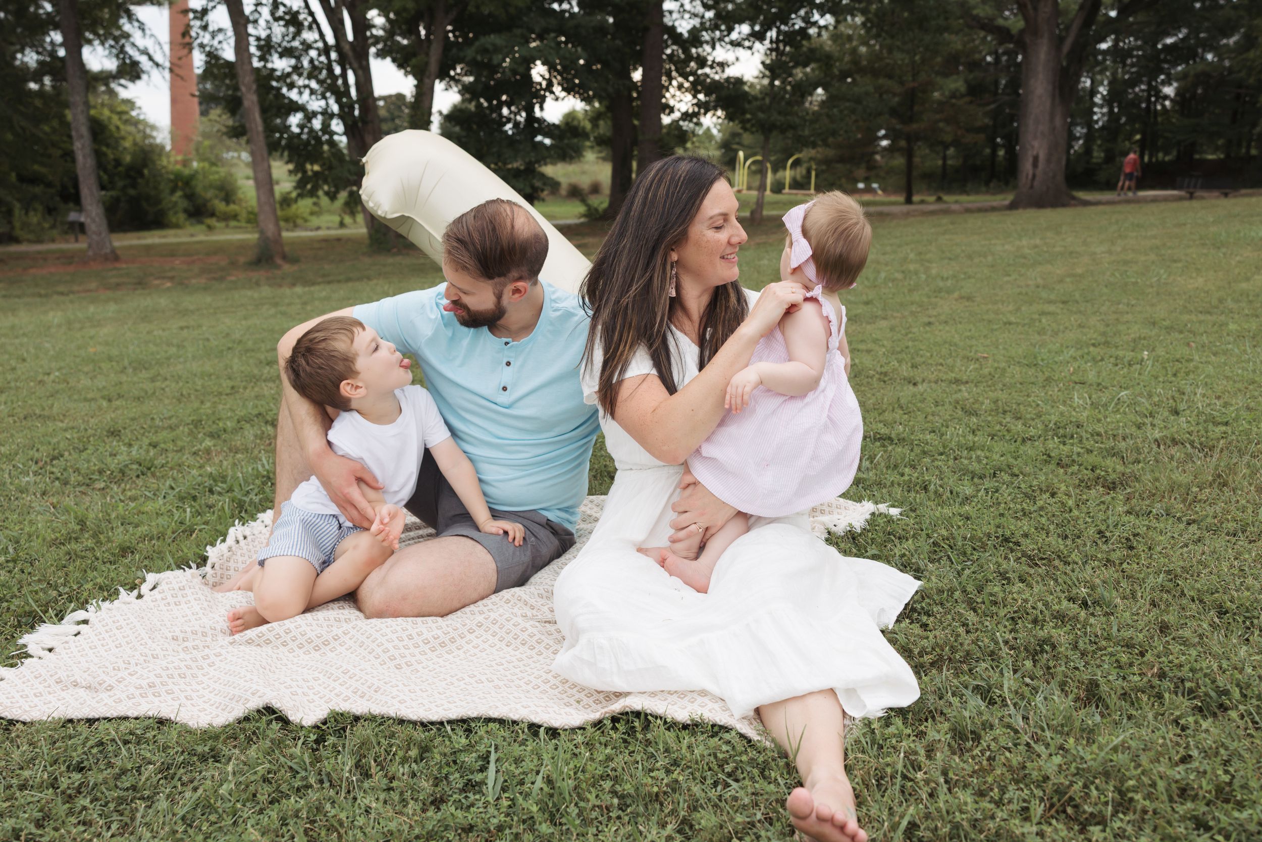 A family sits together on a white blanket enjoying a sunny afternoon in a grassy park.