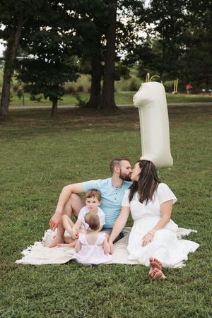 A couple and their baby relax on a white blanket in a grassy park on a sunny afternoon.