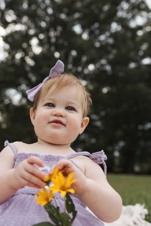 Baby in lavender dress with matching bow holds bright yellow flowers outdoors during summer photoshoot.