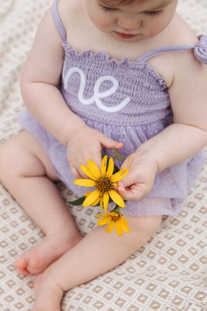 Small baby in purple smocked dress gently holds bright yellow Black-Eyed Susan flowers while sitting on patterned blanket.