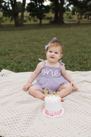 Baby in purple dress sits on white blanket outdoors with small white birthday cake for first birthday celebration.