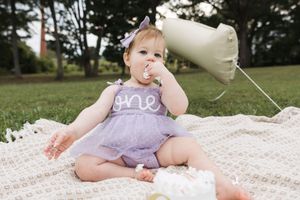 A baby wearing a purple dress sits on a white blanket outdoors playing with balloons.