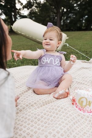 Baby in purple tulle dress sits on blanket during outdoor birthday photoshoot with milestone decorations and balloons.