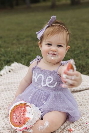 Small child in purple dress sits on blanket eating cake during first birthday photoshoot outdoors.