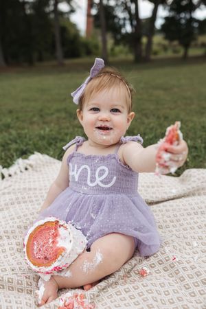 Baby in purple dress sitting on blanket with smashed watermelon enjoying first birthday cake smash photo session.