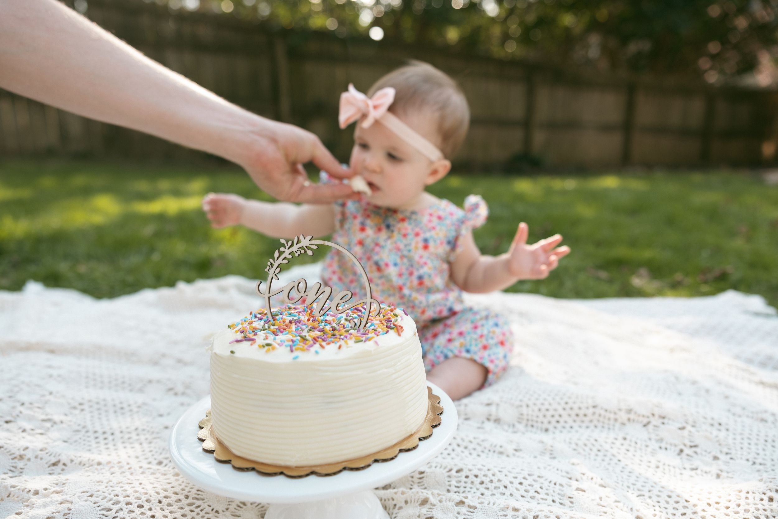 Baby girl in floral outfit sits on white blanket outdoors reaching toward a white birthday smash cake with sprinkles.