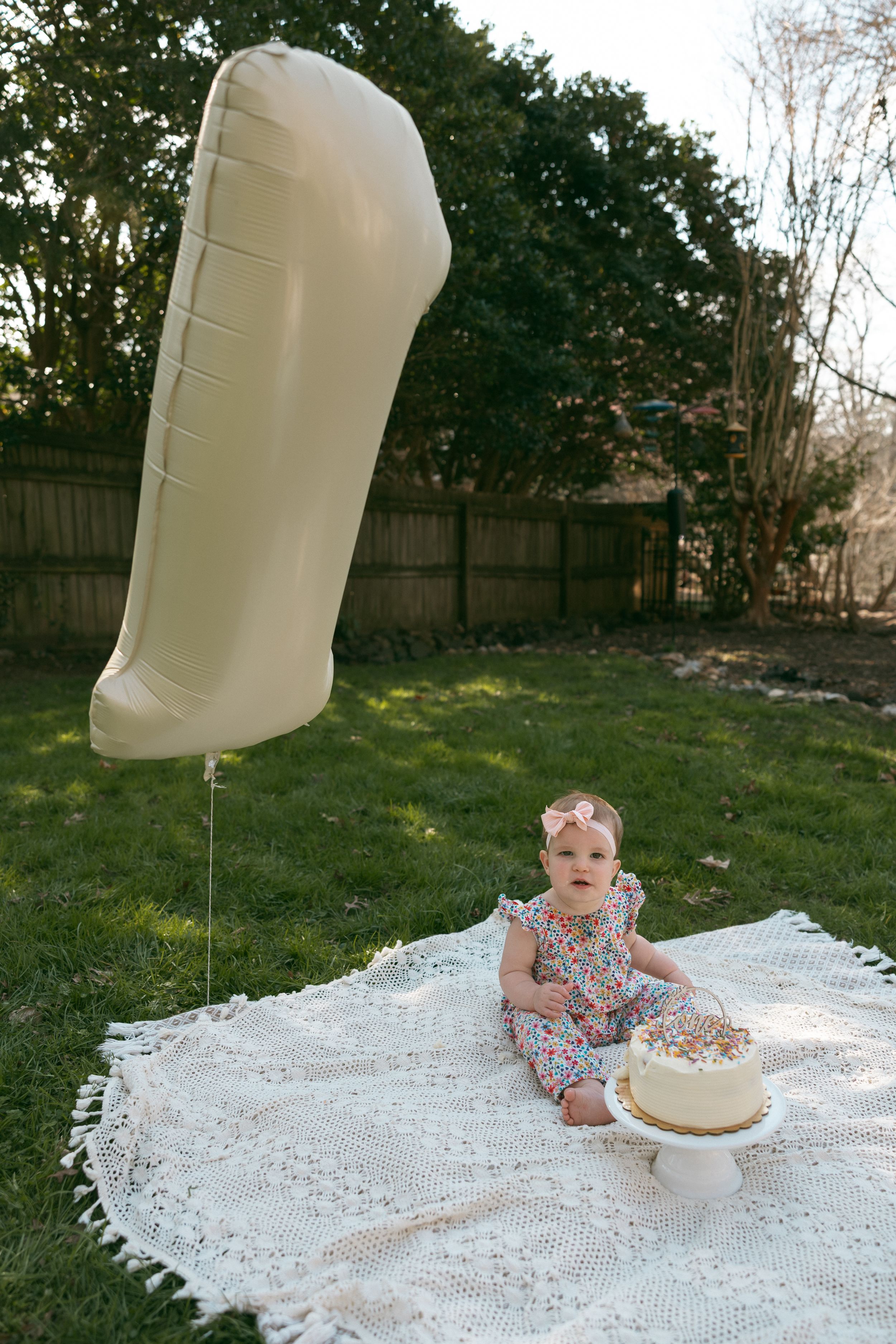 Baby girl in floral dress sits on white blanket outdoors with number 1 balloon and first birthday cake.