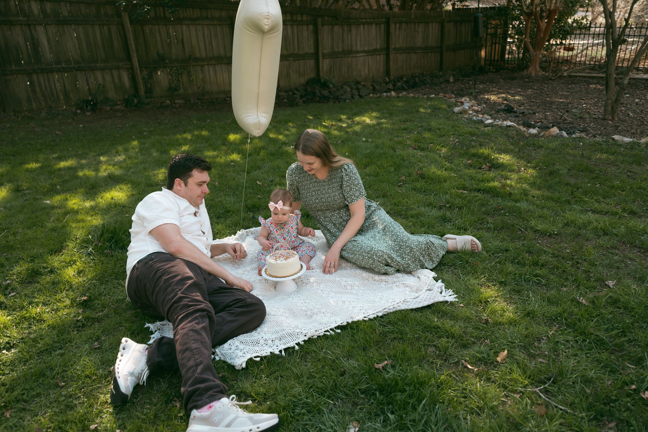 Family celebrating baby's first birthday on a blanket in a backyard with cake and a balloon.