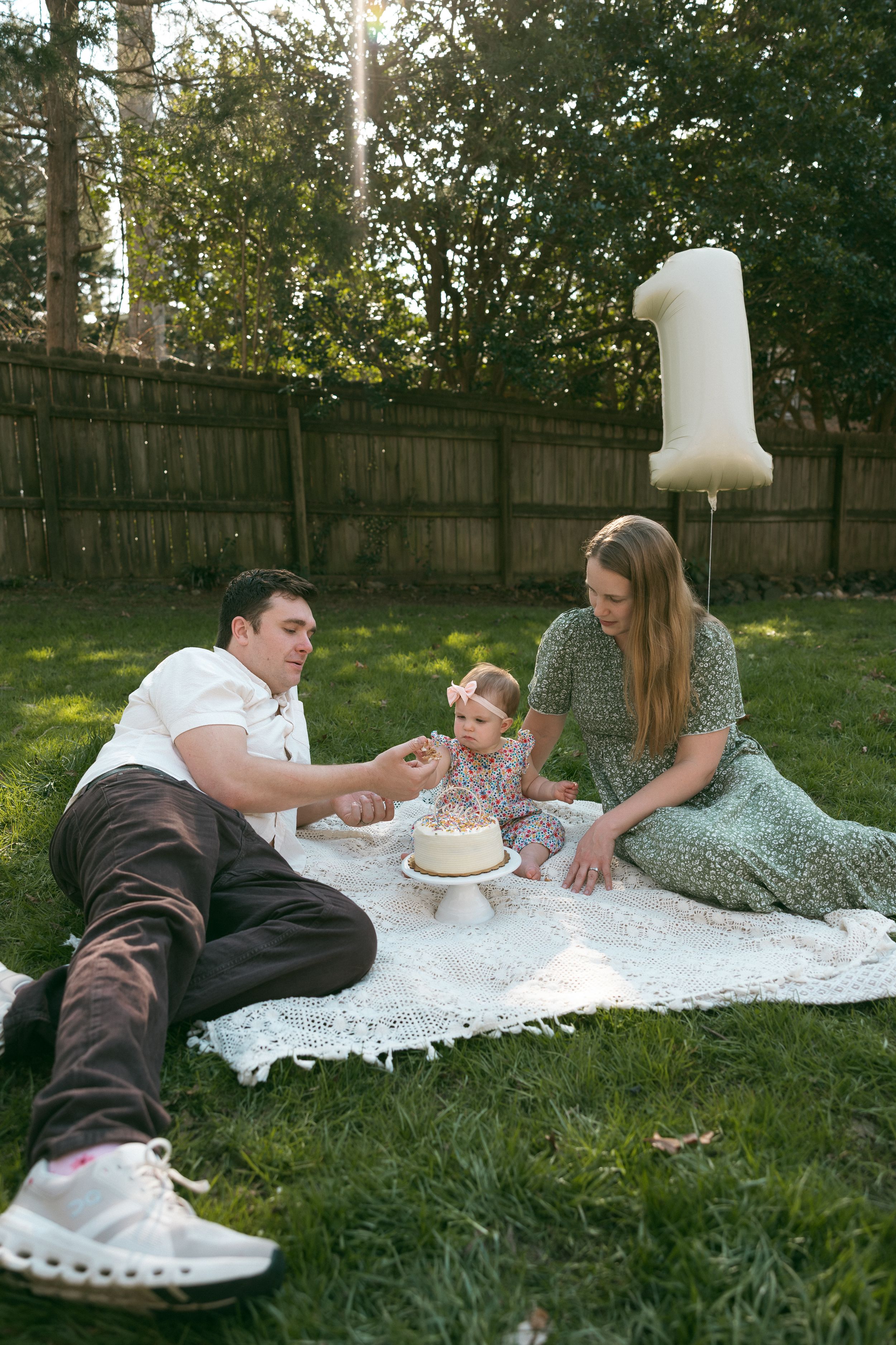 Family celebrating baby's first birthday on a blanket in backyard with white cake and number one balloon.