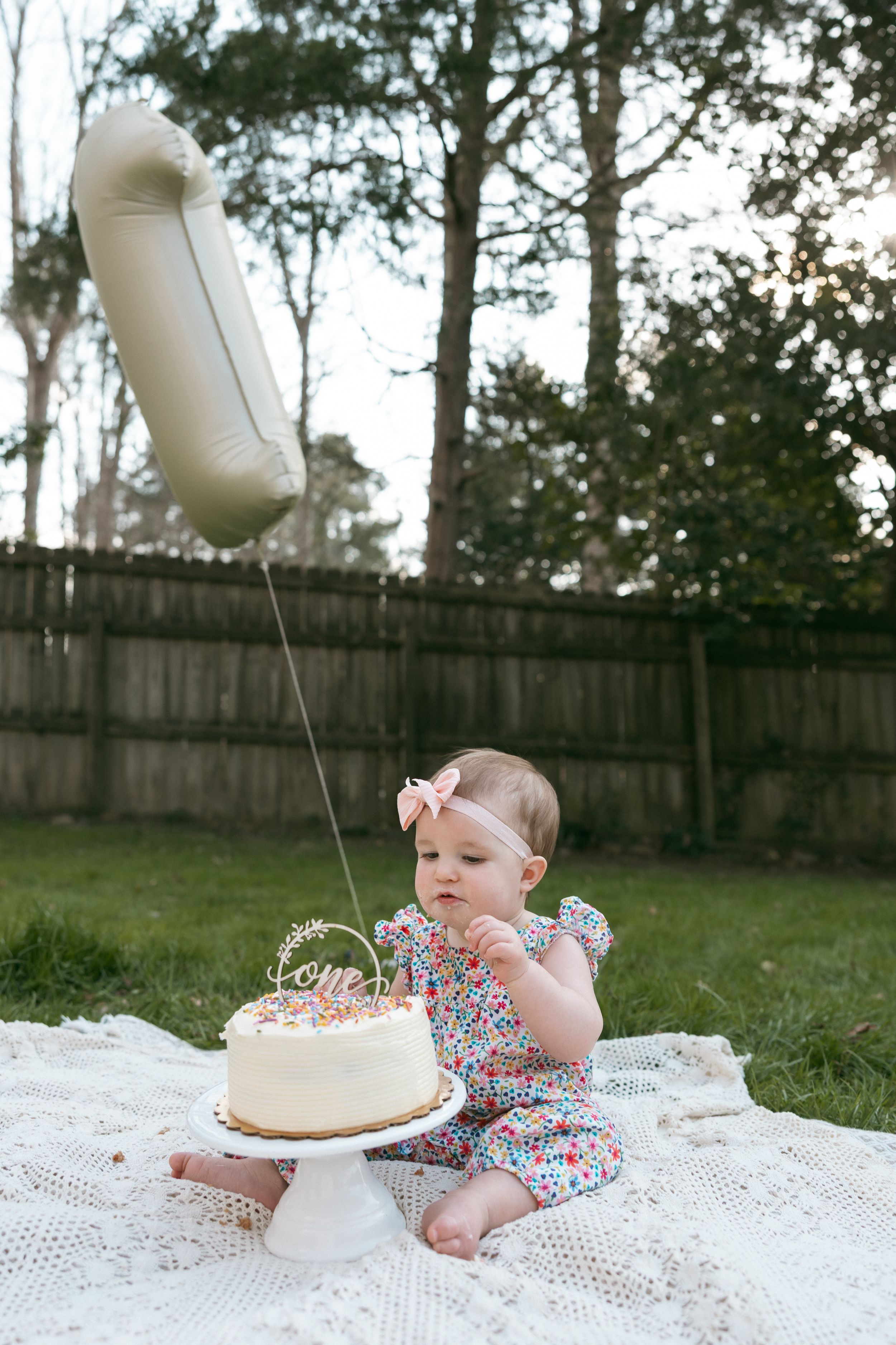 Baby girl in floral dress sits on blanket outdoors touching first birthday cake with number one balloon.