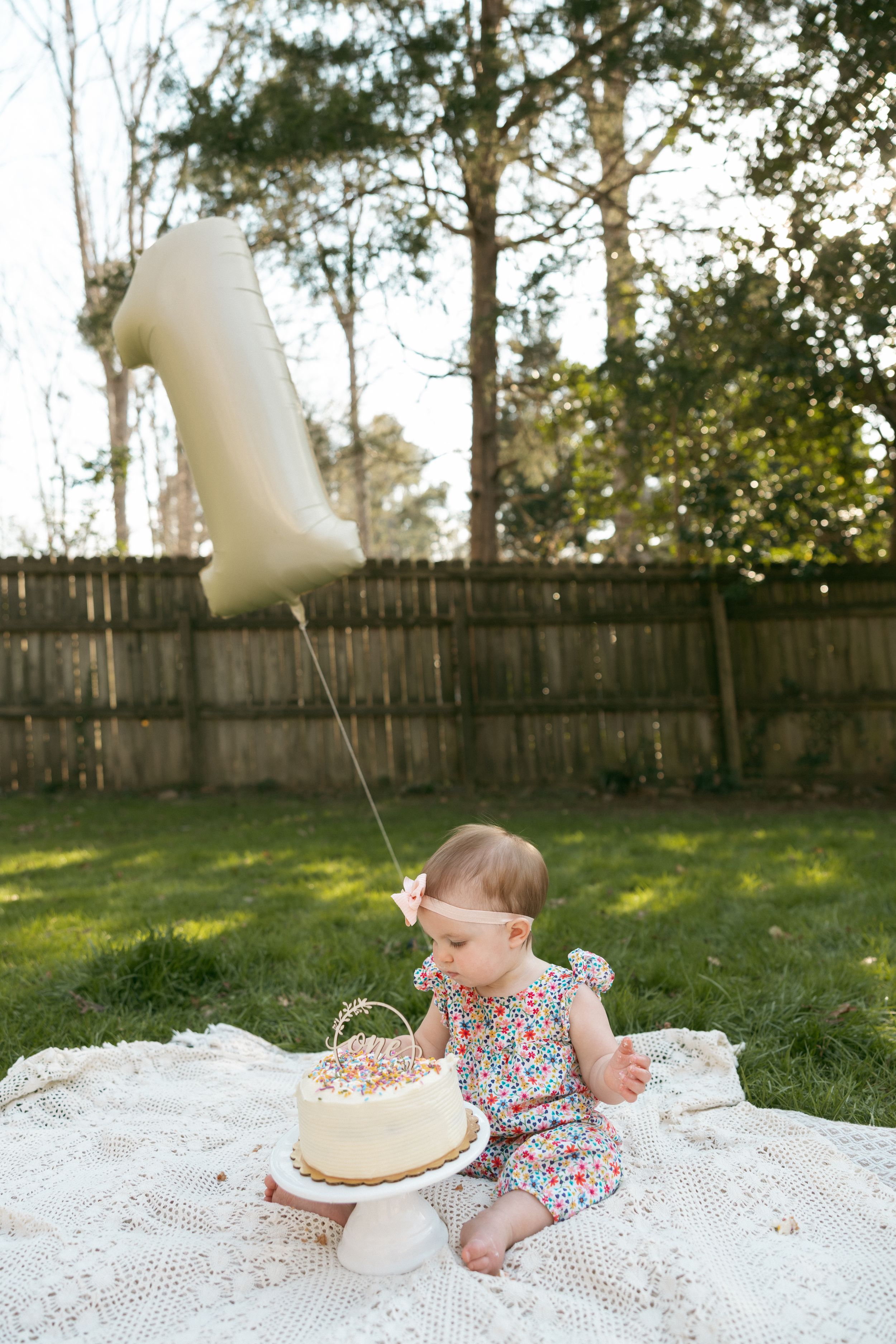 Baby girl celebrates first birthday outdoors with white cake and number one balloon on blanket in sunny backyard.